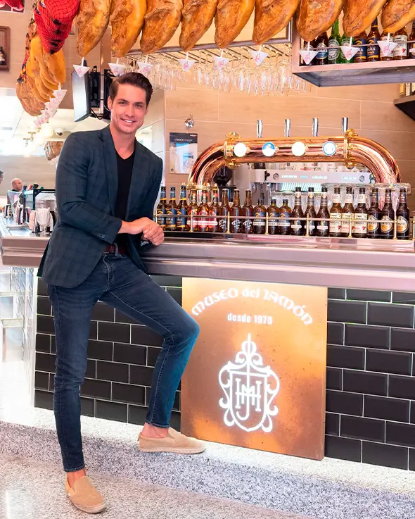 Luis Alfonso Muñoz, CEO del Museo del Jamón, posando en la barra del restaurante con una selección de jamones colgados en el fondo.